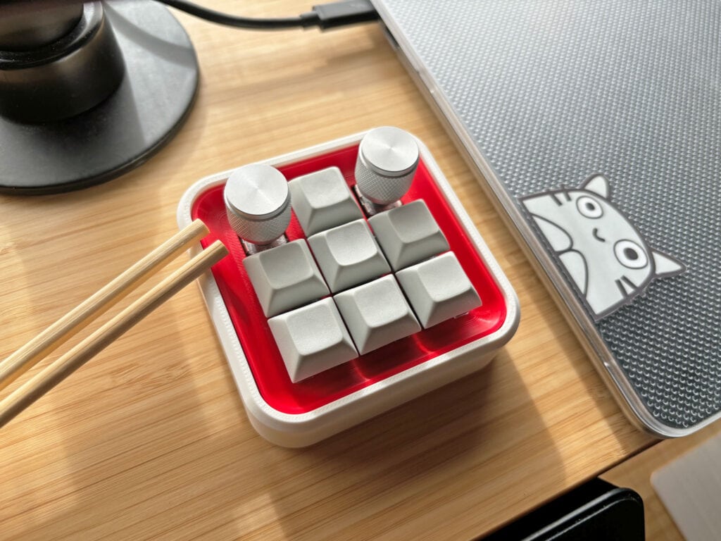 Banchan macropad with a red plate and white case, sitting next to a laptop. A pair of chopsticks are resting on its edge.